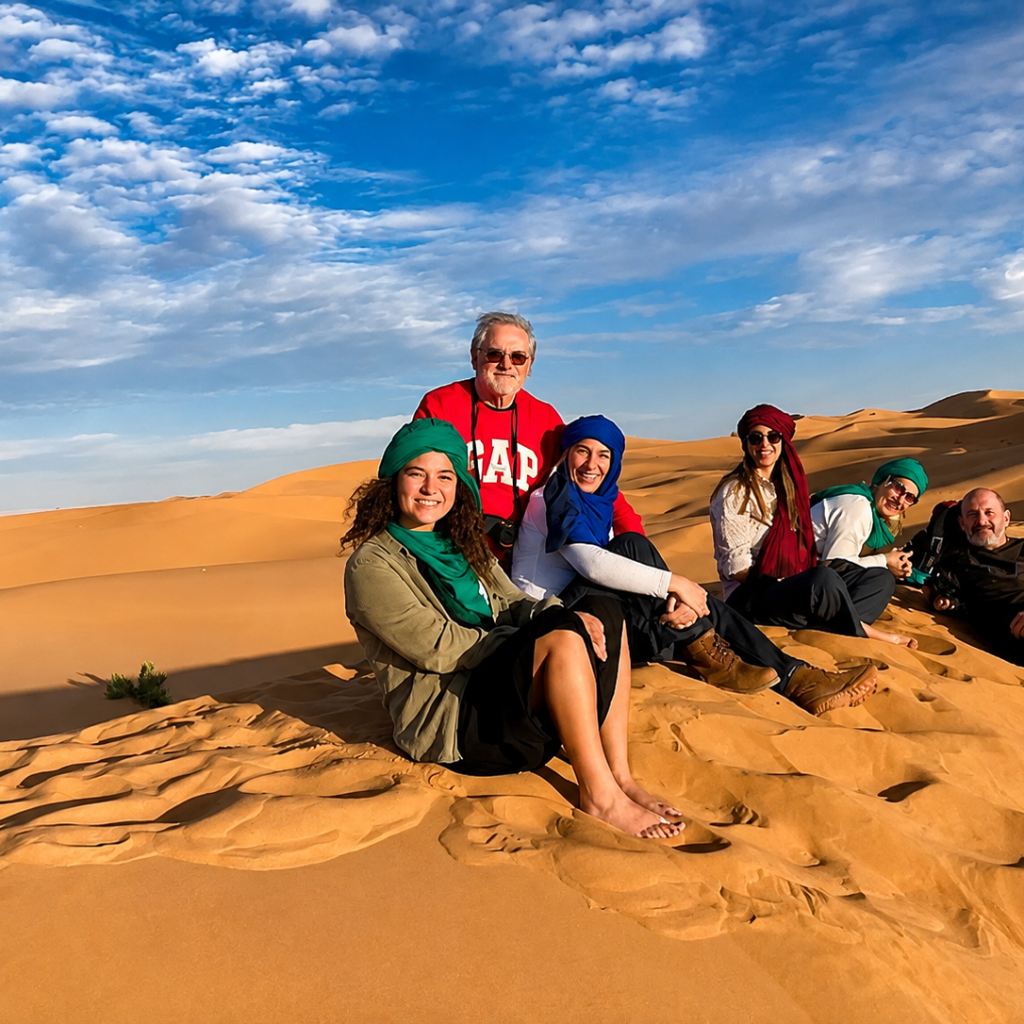 Six travelers sitting together on golden sand dunes in the Sahara Desert under a blue, partly cloudy sky.